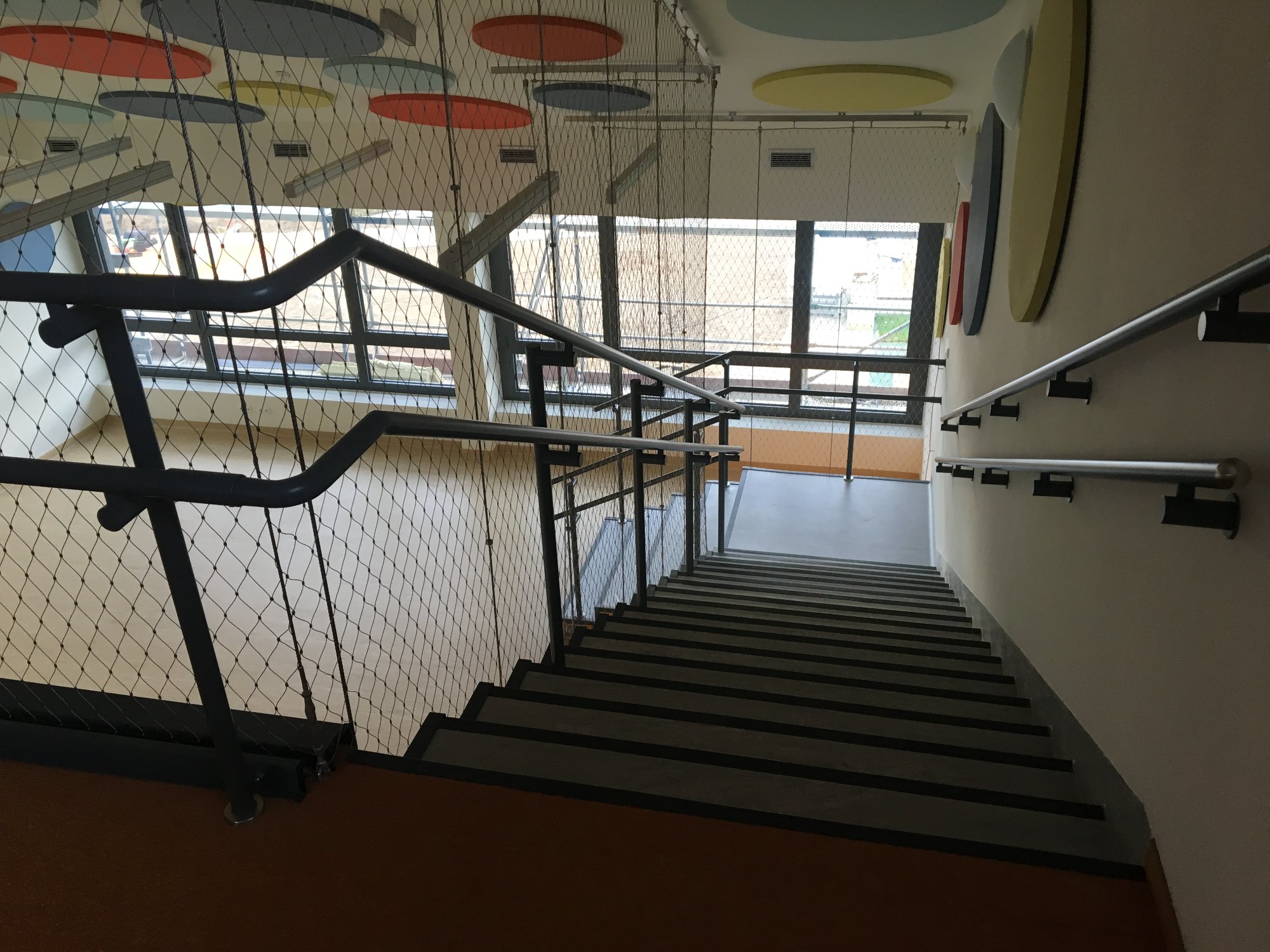 Wnętrze klatki schodowej z widokiem z góry, zabezpieczonej siatką, z kolorowymi okręgami dekoracyjnymi na suficie i ścianie.