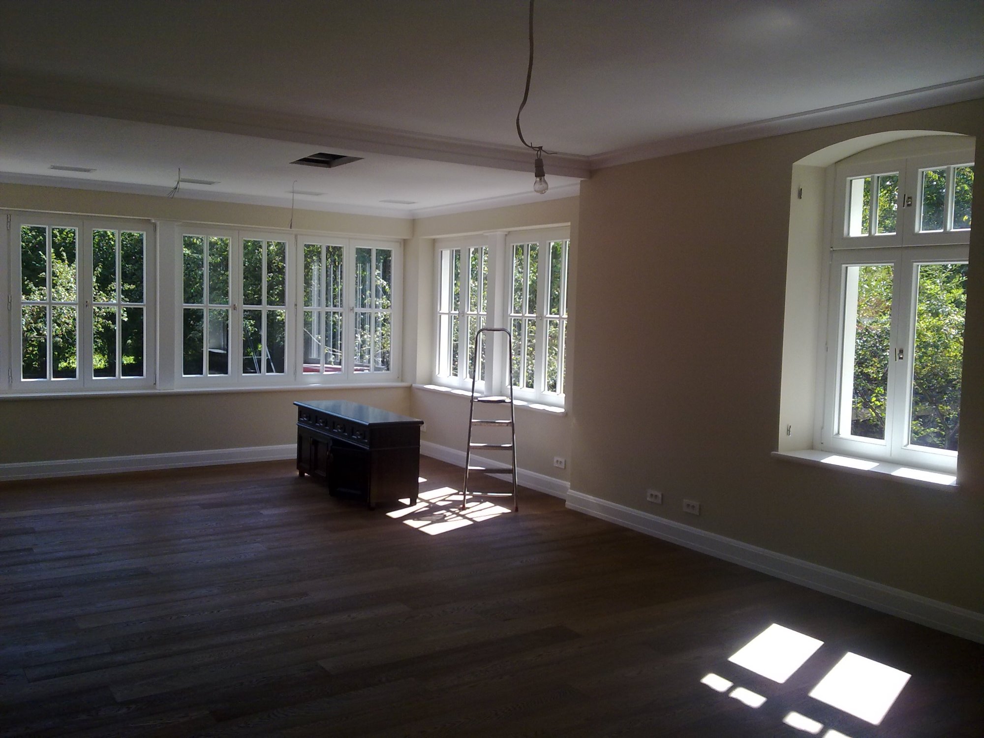 Pusty pokój z drewnianą podłogą, świeżo pomalowanymi ścianami w odcieniach beżu, białymi oknami z widokiem na zieleń, metalową drabiną i ciemną komodą. Na suficie widoczne niedokończone elementy...