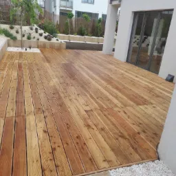 Drewniany taras z desek w odcieniach brązu przylega do białego budynku z dużymi przesuwnymi drzwiami, otoczony białymi kamykami i zielenią.