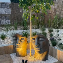 Oświetlony ogród z dwoma rzeźbami głowy, drzewem kulistym, białym żwirem i drewnianym tarasem w tle bambusowy płot.