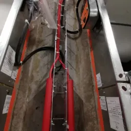 Metalowa drabinka kablowa z zamontowanymi przewodami elektrycznymi w czerwonej izolacji, zabezpieczonymi uchwytami, oraz skrzynka kontrolna zamontowana na boku drabinki, widoczne etykiety...