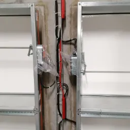 Widok z bliska instalacji elektrycznej w prowadnicy kablowej, zamontowanej pomiędzy dwoma segmentami bramy garażowej z napędem, zabezpieczonym folią ochronną.