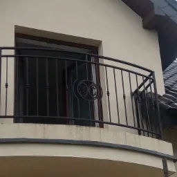 Balkon z czarną metalową balustradą z ozdobnym elementem na tle beżowej elewacji domu z ciemnym dachem.