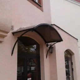 Zadaszenie nad wejściem do budynku z metalową konstrukcją i przydymionym brązowym poliwęglanem, wsparte na ozdobnych, kutych wspornikach z lampą, na tle elewacji w kolorze pudrowego różu.