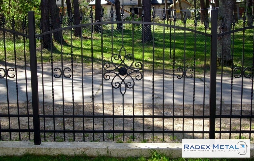 Czarne, metalowe ogrodzenie z dekoracyjnymi elementami w kształcie spirali i rozet, zamontowane na betonowej podmurówce, z widocznym logo Radex Metal w prawym dolnym rogu.