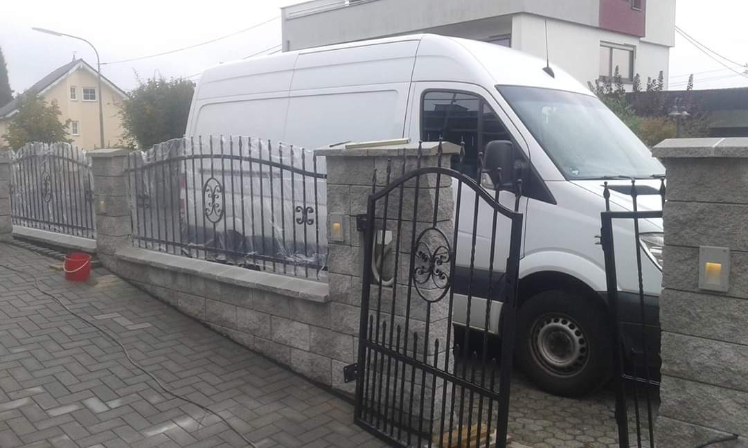 Nowoczesne ogrodzenie z metalowymi przęsłami i kamiennymi słupkami, częściowo zakryte przez białego dostawczaka, z otwartą furtką i widocznym fragmentem posesji.