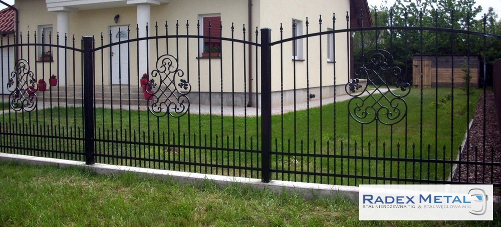 Czarne, metalowe przęsła ogrodzeniowe z ozdobnymi elementami na tle domu z czerwonymi doniczkami na parapecie.