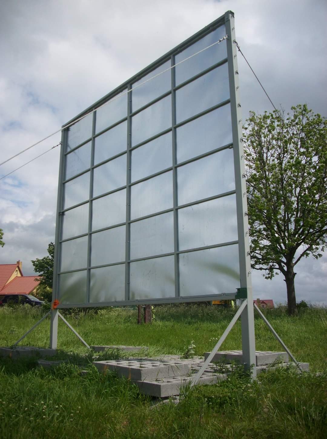 Dźwiękochłonna konstrukcja stalowa w Słupsku, z podziałem na kwadratowe panele, zabezpieczona linkami, na betonowej podstawie, w otoczeniu zieleni i drzew.
