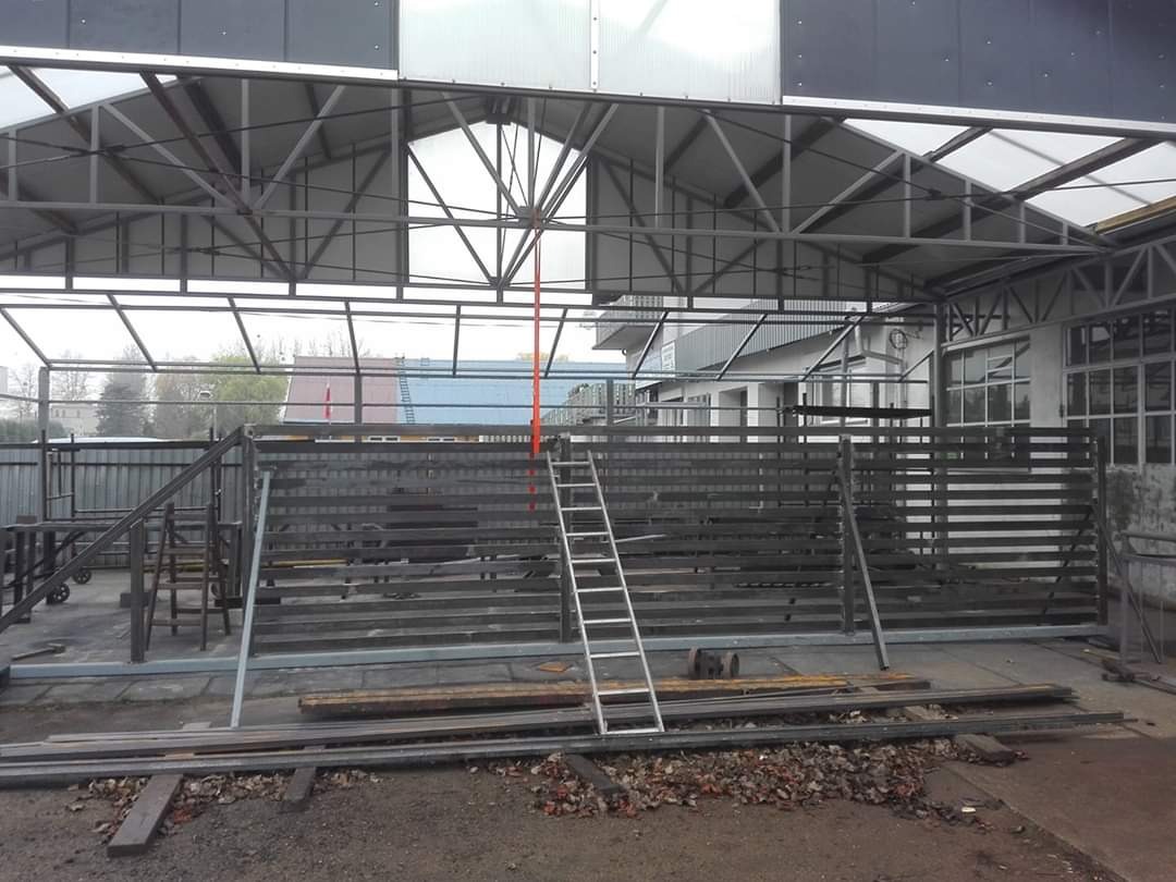 Konstrukcja metalowa bramy przesuwnej w trakcie montażu, widoczna drabina oparta o konstrukcję, elementy metalowe na ziemi, zadaszenie nad miejscem pracy.