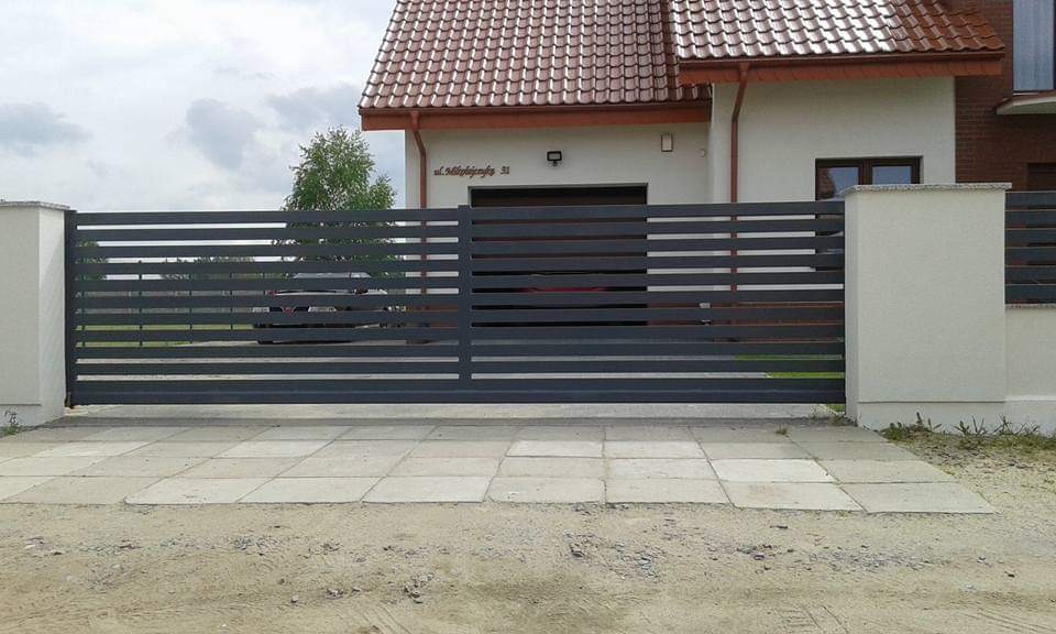 Nowoczesna, przesuwna brama wjazdowa z poziomych, metalowych profili pomalowanych na grafitowo, zamontowana na posesji z jasnym domem i kostką brukową na podjeździe.