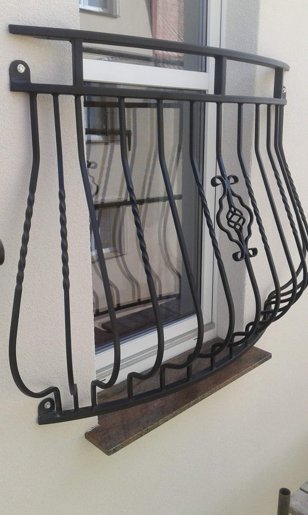 Czarna, metalowa balustrada okienna z ozdobnym elementem, zamontowana na jasnej elewacji nad kamiennym parapetem.