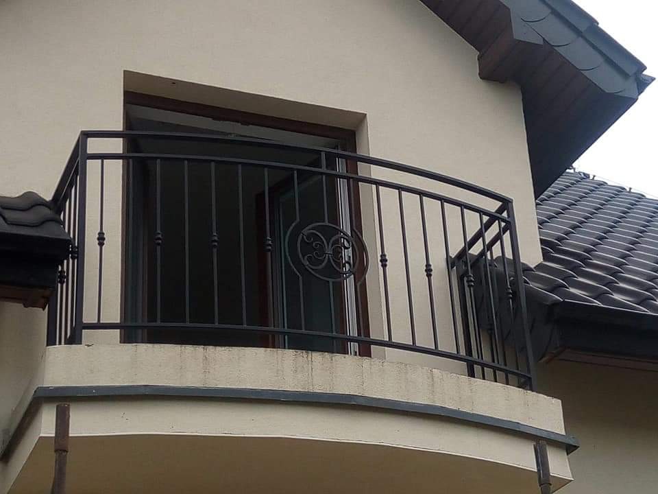 Balkon z czarną metalową balustradą z ozdobnym elementem na tle beżowej elewacji domu z ciemnym dachem.