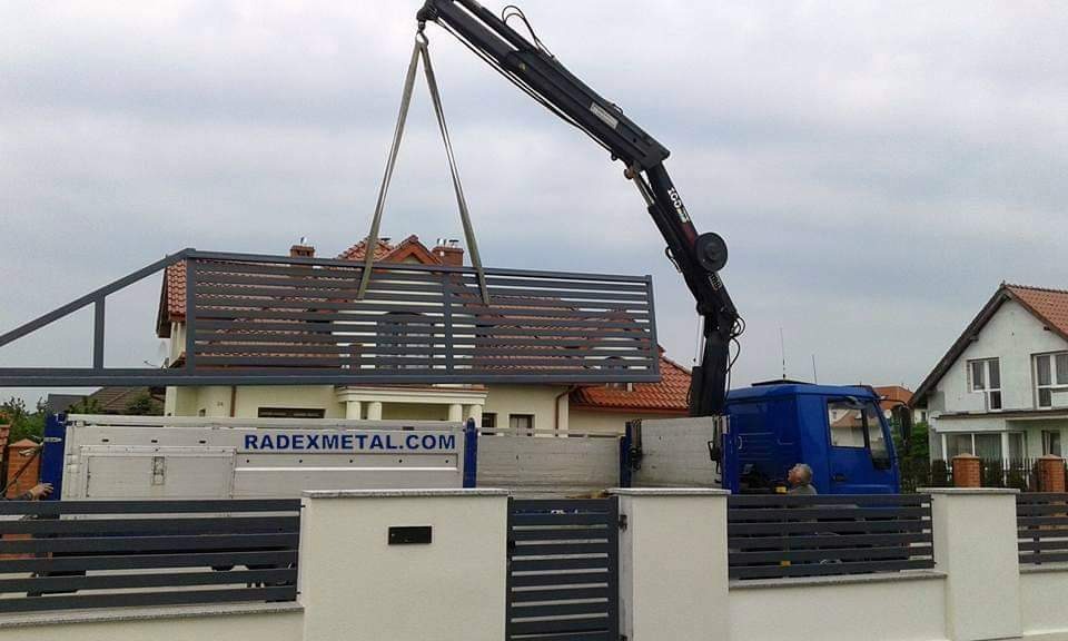 Montaż przęsła ogrodzenia metalowego z poziomych profili przez dźwig samochodowy Radexmetal.com na tle zabudowy jednorodzinnej.
