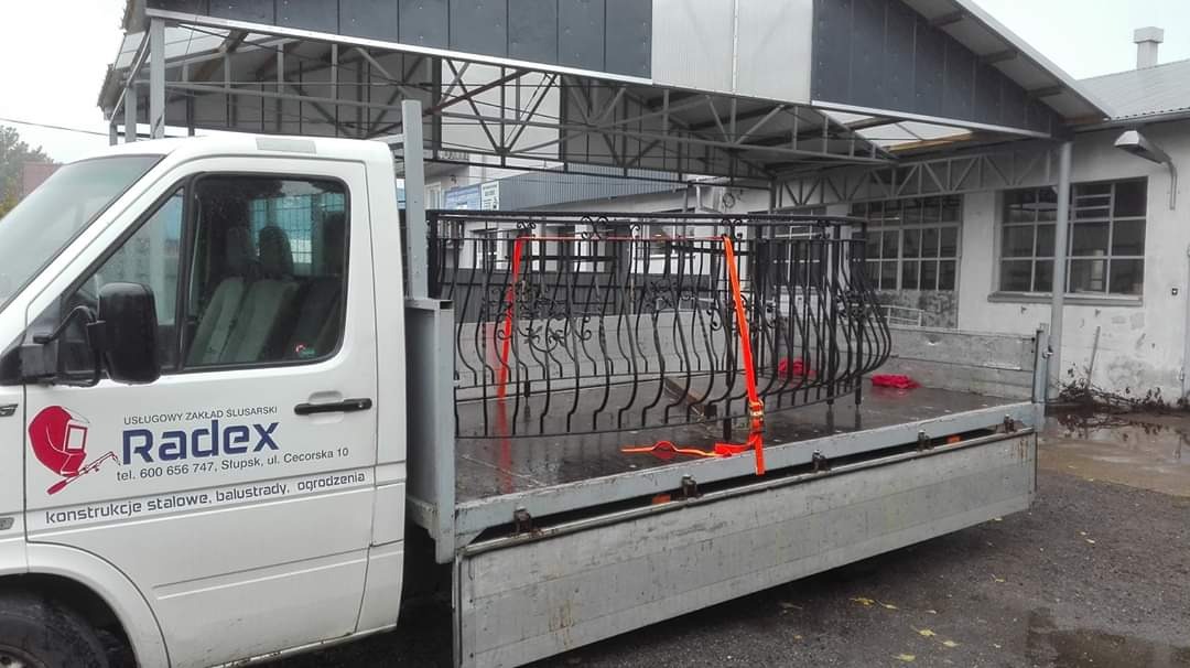 Transport metalowego balkonu z ozdobnymi elementami w samochodzie dostawczym firmy Radex z Słupska, zabezpieczonego pomarańczowymi pasami.