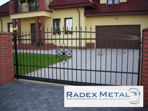 Czarna, metalowa brama przesuwna z dekoracyjnym ornamentem na tle domu jednorodzinnego z garażem. Brama zamontowana pomiędzy dwoma czerwonymi, ceglanymi słupkami.