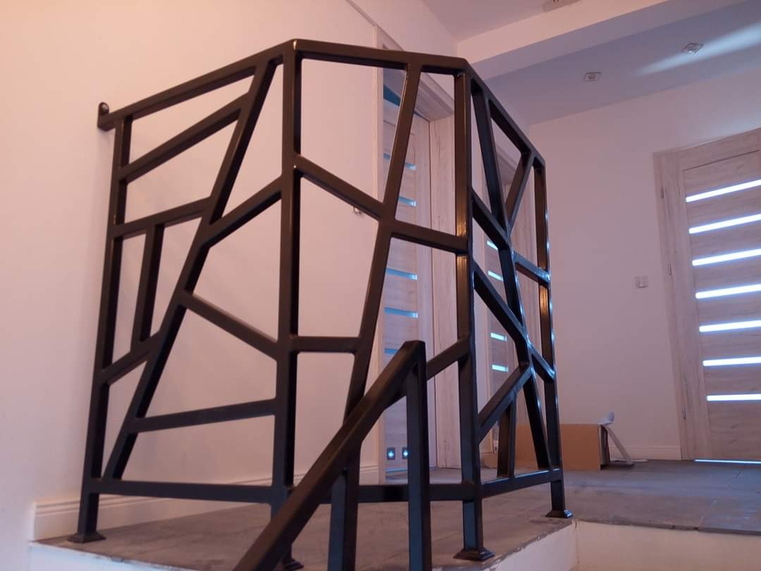 Nowoczesna, czarna balustrada metalowa o geometrycznym wzorze, zamontowana na schodach w jasnym wnętrzu z widocznymi drzwiami.