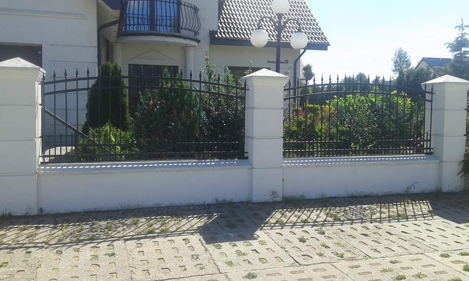 Czarne, metalowe ogrodzenie z ozdobnymi grotami, osadzone na niskim, białym murku z kwadratowymi słupkami, przed posesją z białym domem i lampą w kształcie kul.