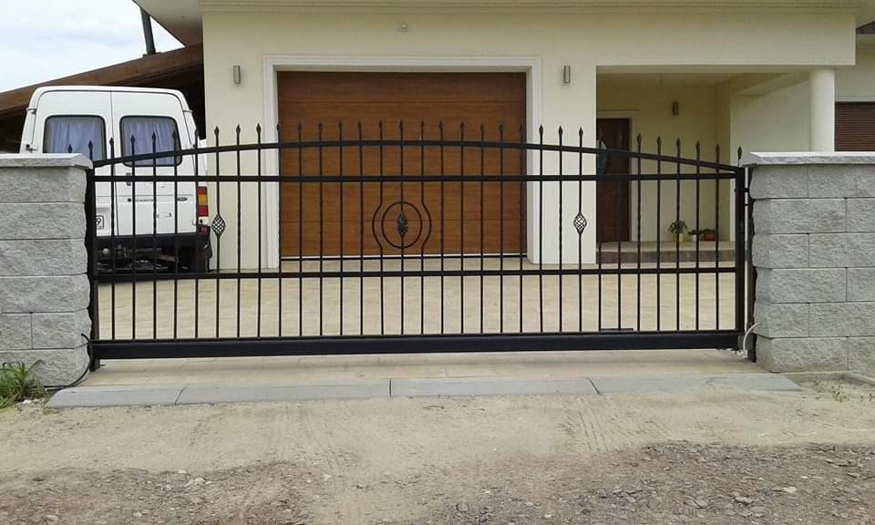 Czarna, metalowa brama przesuwna z ozdobnymi elementami, zamontowana między dwoma słupkami z szarego kamienia, na tle budynku z brązową bramą garażową i białym vanem.