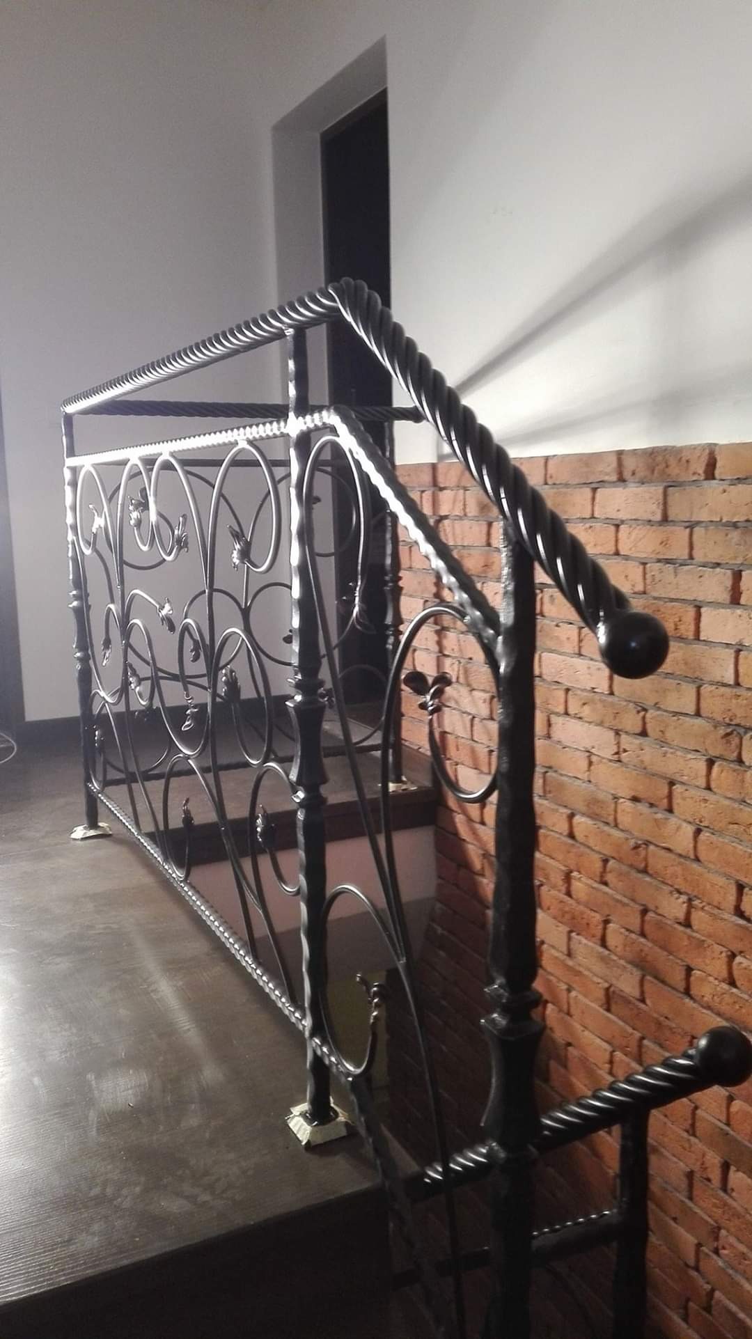 Kute, czarne balustrady schodowe z motywem roślinnym, zamontowane przy ścianie z czerwonej cegły.