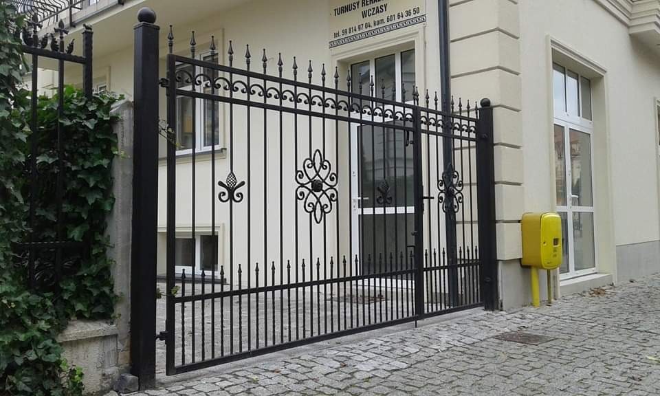 Czarna, metalowa brama wjazdowa z ozdobnymi elementami, osadzona pomiędzy murowanymi słupkami, na tle budynku z żółtą skrzynką elektryczną i brukowaną nawierzchnią