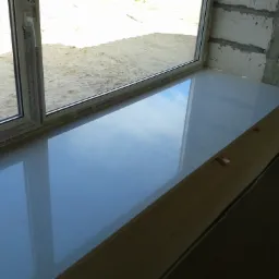 Nowo zamontowane okno z osadzonym białym parapetem, widok z wnętrza budynku na piaszczyste podłoże na zewnątrz, okno marki Salamander z widocznymi napisami na ramie.