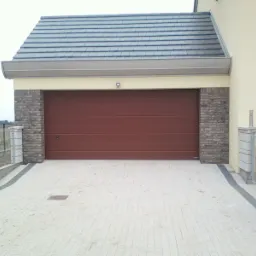 Brązowa brama garażowa segmentowa z poziomymi przetłoczeniami, otoczona ceglanymi filarami, z widocznym fragmentem posesji i domu z szarą dachówką.