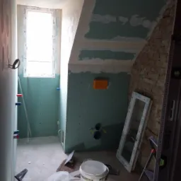 Łazienka w trakcie prac wykończeniowych: ściany pokryte płytami kartonowo-gipsowymi, widoczne instalacje wodne, okno, drabina, poziomica Stanley Fatmax oraz wiadro z masą szpachlową na podłodze.