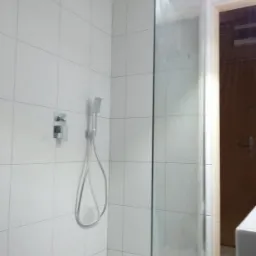 Nowoczesna kabina prysznicowa z deszczownicą i panelem sterującym, wyłożona białymi, kwadratowymi płytkami, widoczna przezroczysta szyba i fragment drzwi w tle.