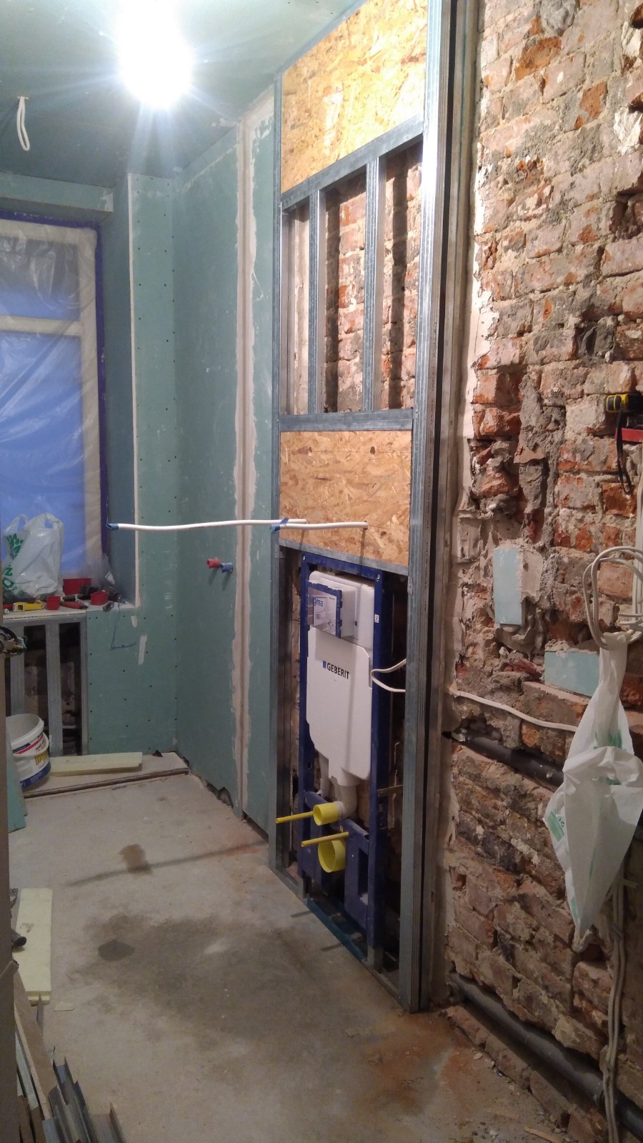 Łazienka w trakcie prac: widoczny stelaż podtynkowy Geberit zamontowany na tle surowej ceglanej ściany, częściowo obudowany płytą OSB i metalowym profilem, ściany pokryte płytami...