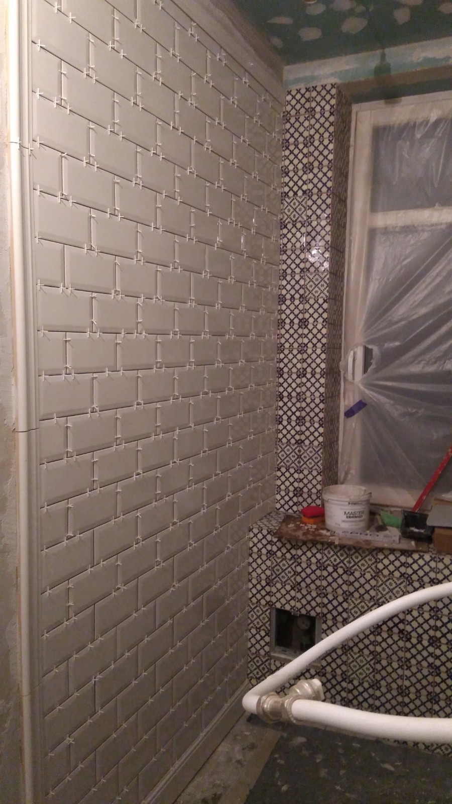 Ściana łazienki w trakcie układania płytek typu metro, z białymi krzyżykami dystansowymi i fragmentem dekoracyjnej mozaiki na sąsiedniej ścianie, okno zakryte folią ochronną.