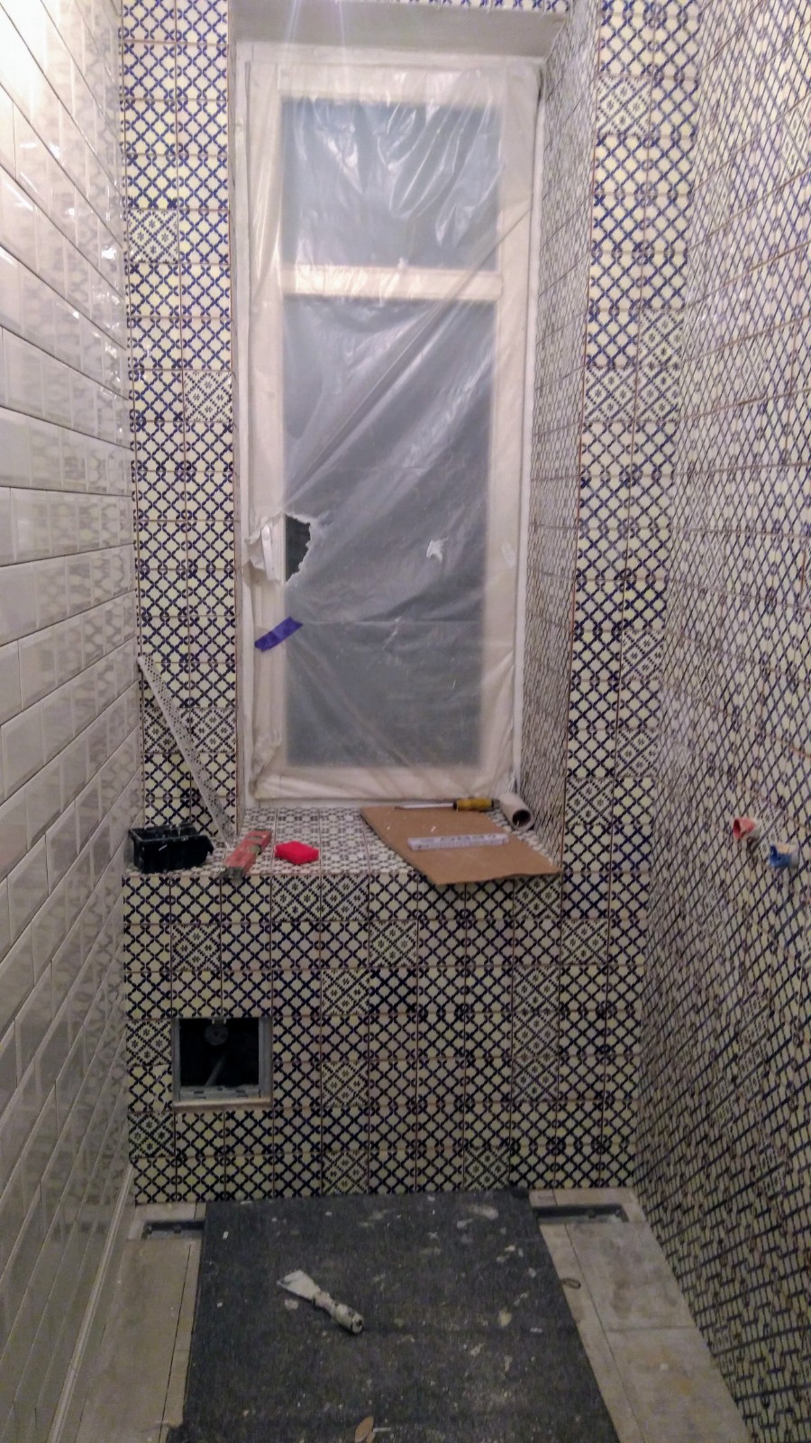 Łazienka w trakcie prac wykończeniowych: częściowo wyłożona białymi i wzorzystymi płytkami, okno zabezpieczone folią, widoczne narzędzia i materiały budowlane, podłoga częściowo zakryta.