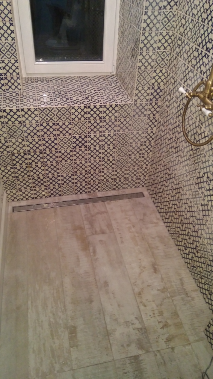 Ukończona kabina prysznicowa z odpływem liniowym, wyłożona płytkami w geometryczne wzory w odcieniach bieli i granatu, z drewnopodobną podłogą i mosiężną armaturą.