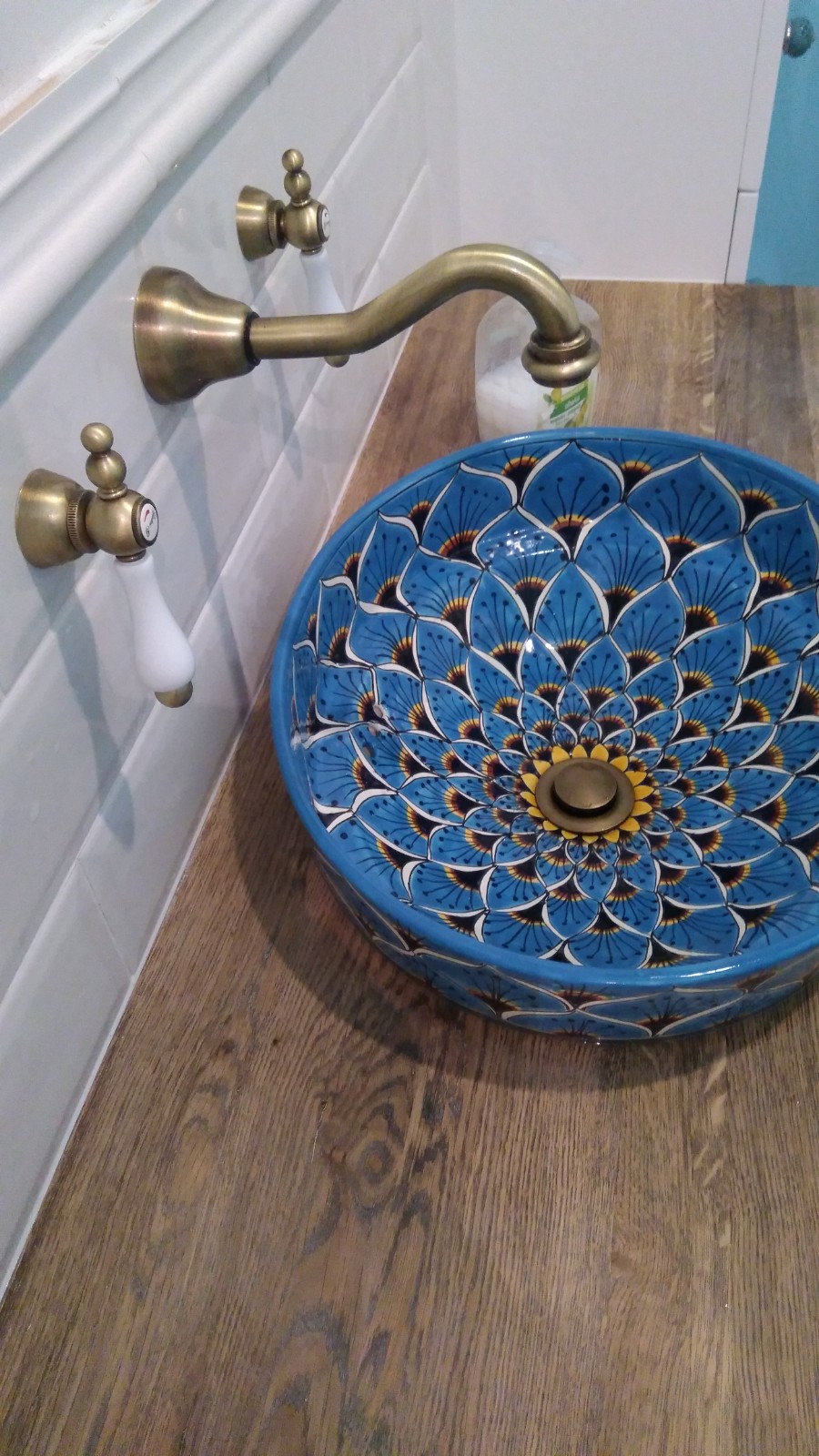 Umywalka ceramiczna w kolorze niebieskim z wzorem kwiatowym, zamontowana na drewnianym blacie, z baterią w stylu retro z białymi kurkami i płytkami ceramicznymi w tle.