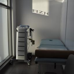Klinika Fizjoterapii Zbigniew Mittelstadt - Jasne wnętrze gabinetu fizjoterapii z aparaturą do zabiegów, łóżkiem rehabilitacyjnym z wałkiem i napisem 'FIZJO' odbitym od okna na ścianie.