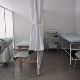 Jasne wnętrze gabinetu z dwoma leżankami rehabilitacyjnymi z jasnym obiciem, szarą aparaturą do krioterapii z przezroczystym blatem, białymi zasłonami i krzesłami na szarej podłodze.