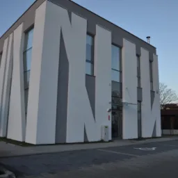 Nowoczesny budynek kliniki z charakterystyczną fasadą w biało-szare pasy, widoczne okna, wejście z zadaszeniem, chodnik z kostki brukowej i oznakowane miejsce parkingowe dla osób niepełnosprawnych.