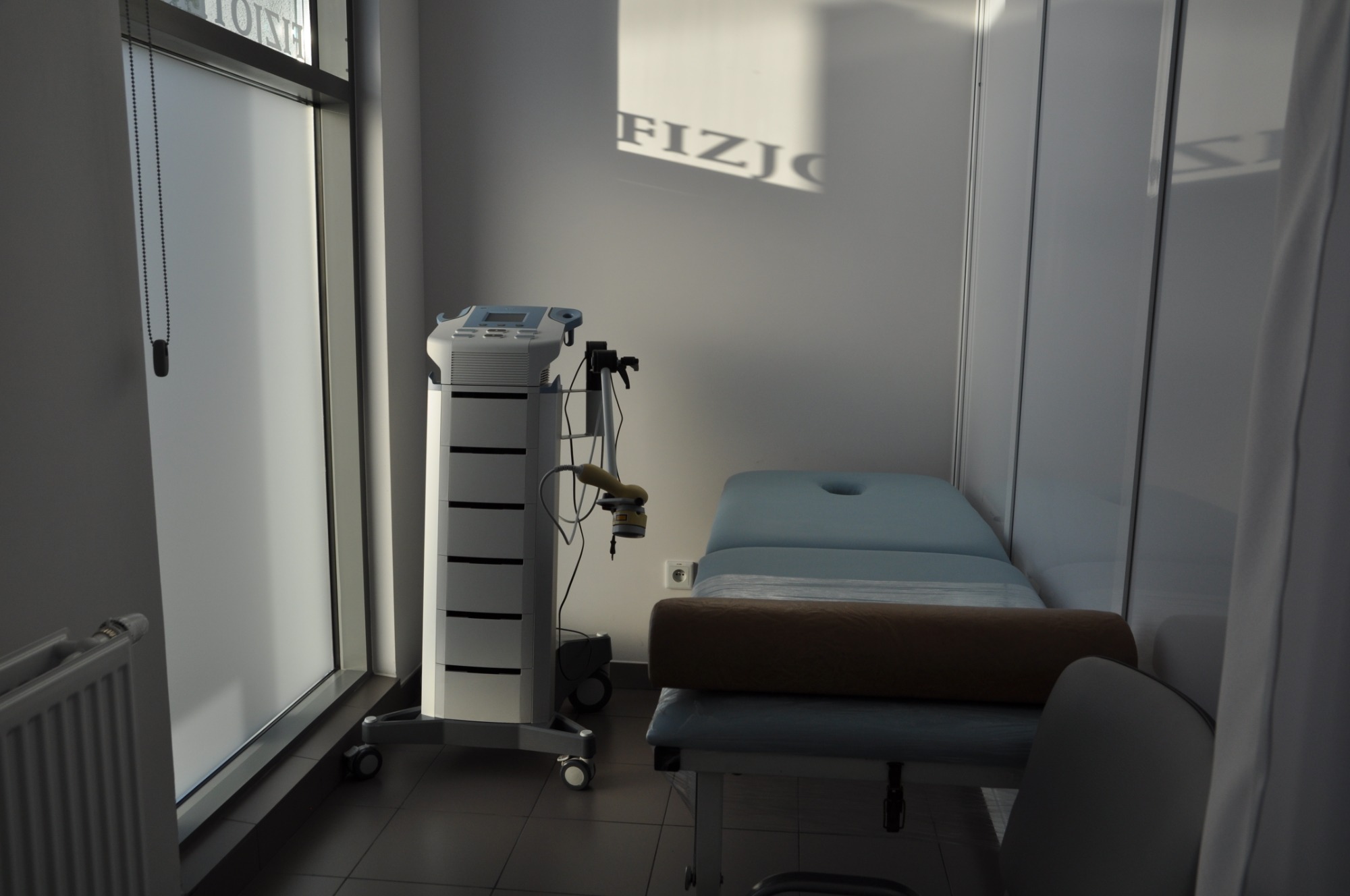 Jasne wnętrze gabinetu fizjoterapii z aparaturą do zabiegów, łóżkiem rehabilitacyjnym z wałkiem i napisem 'FIZJO' odbitym od okna na ścianie.