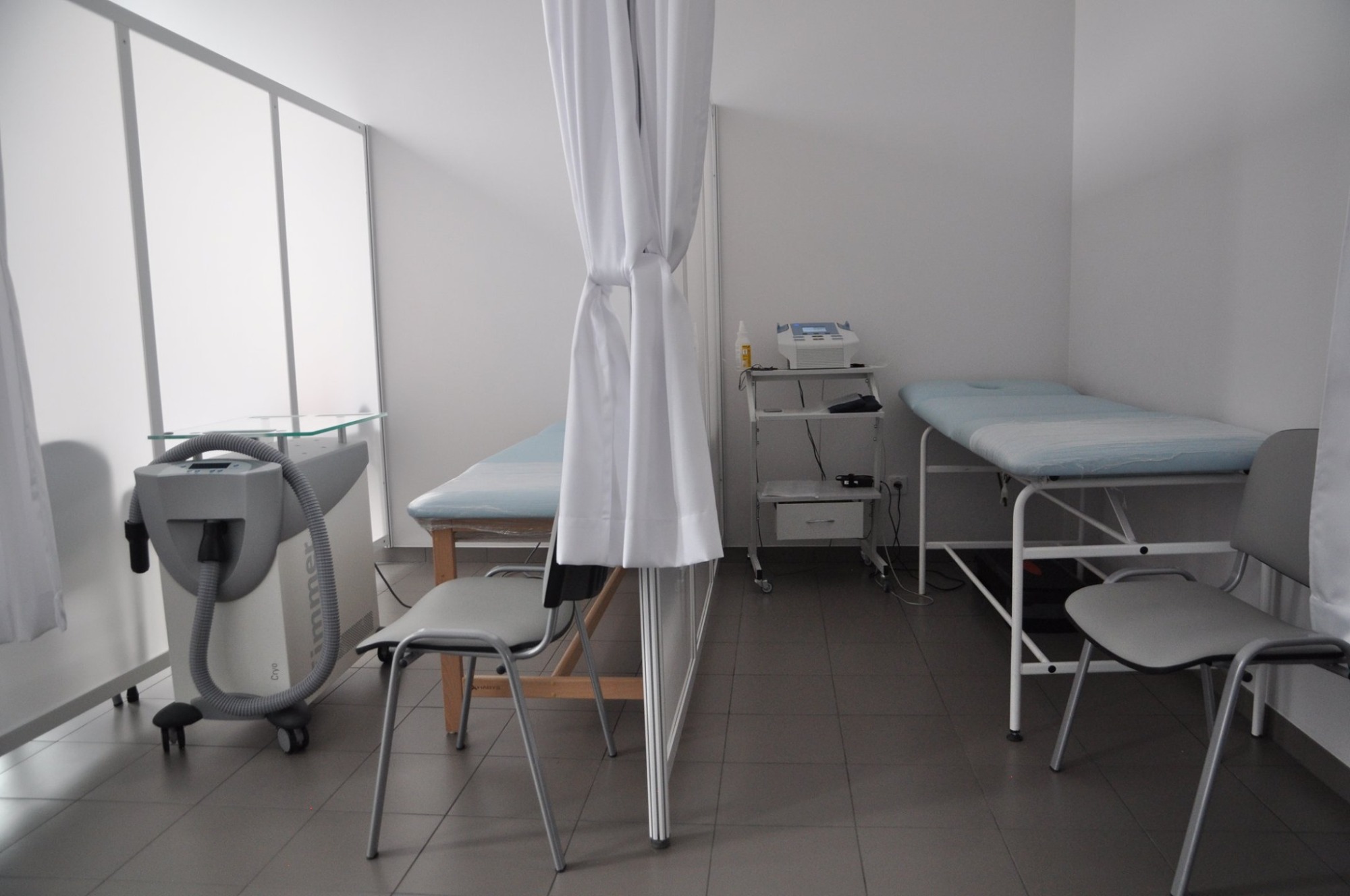Jasne wnętrze gabinetu z dwoma leżankami rehabilitacyjnymi z jasnym obiciem, szarą aparaturą do krioterapii z przezroczystym blatem, białymi zasłonami i krzesłami na szarej podłodze.
