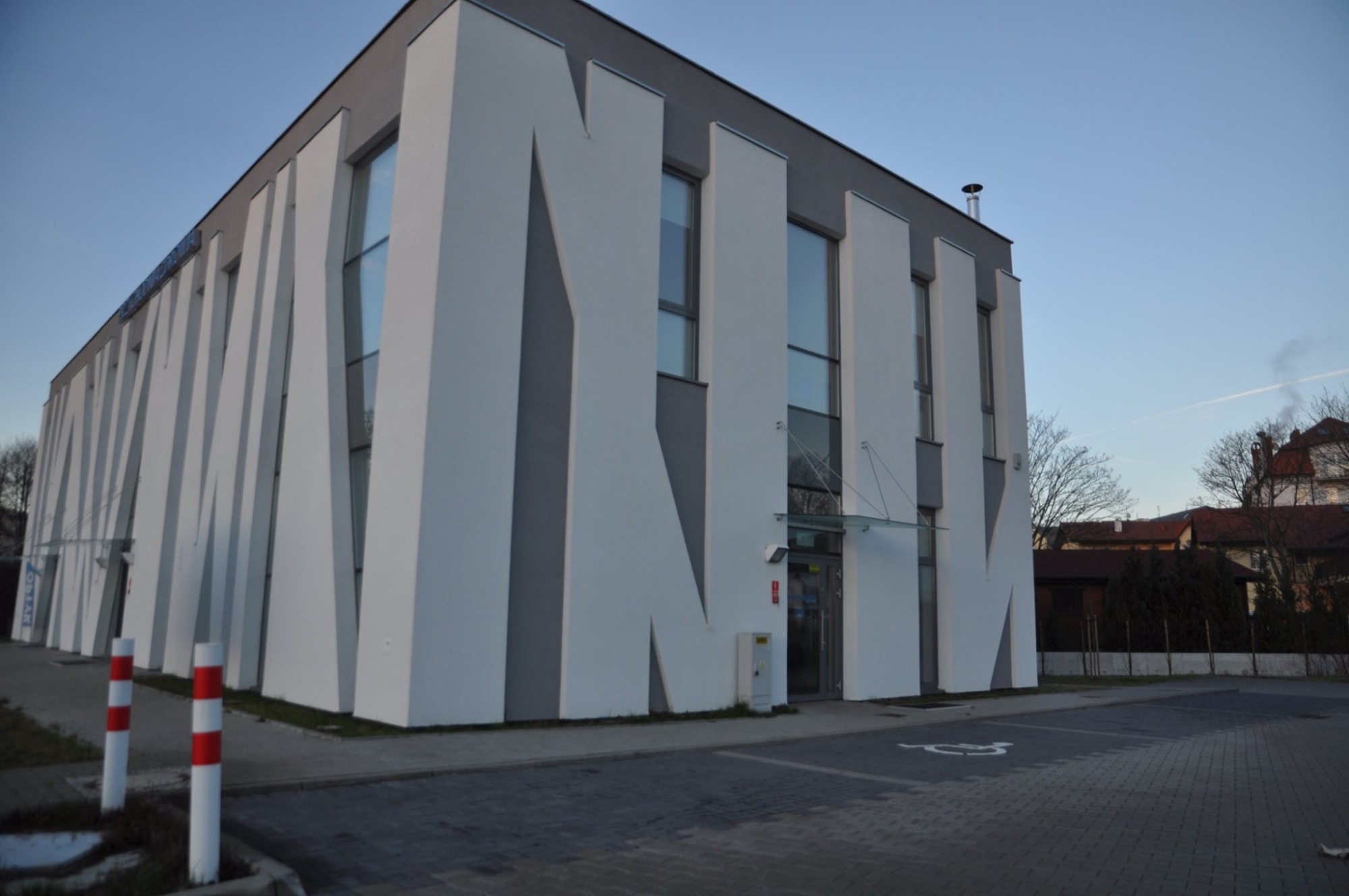 Nowoczesny budynek kliniki z charakterystyczną fasadą w biało-szare pasy, widoczne okna, wejście z zadaszeniem, chodnik z kostki brukowej i oznakowane miejsce parkingowe dla osób niepełnosprawnych.