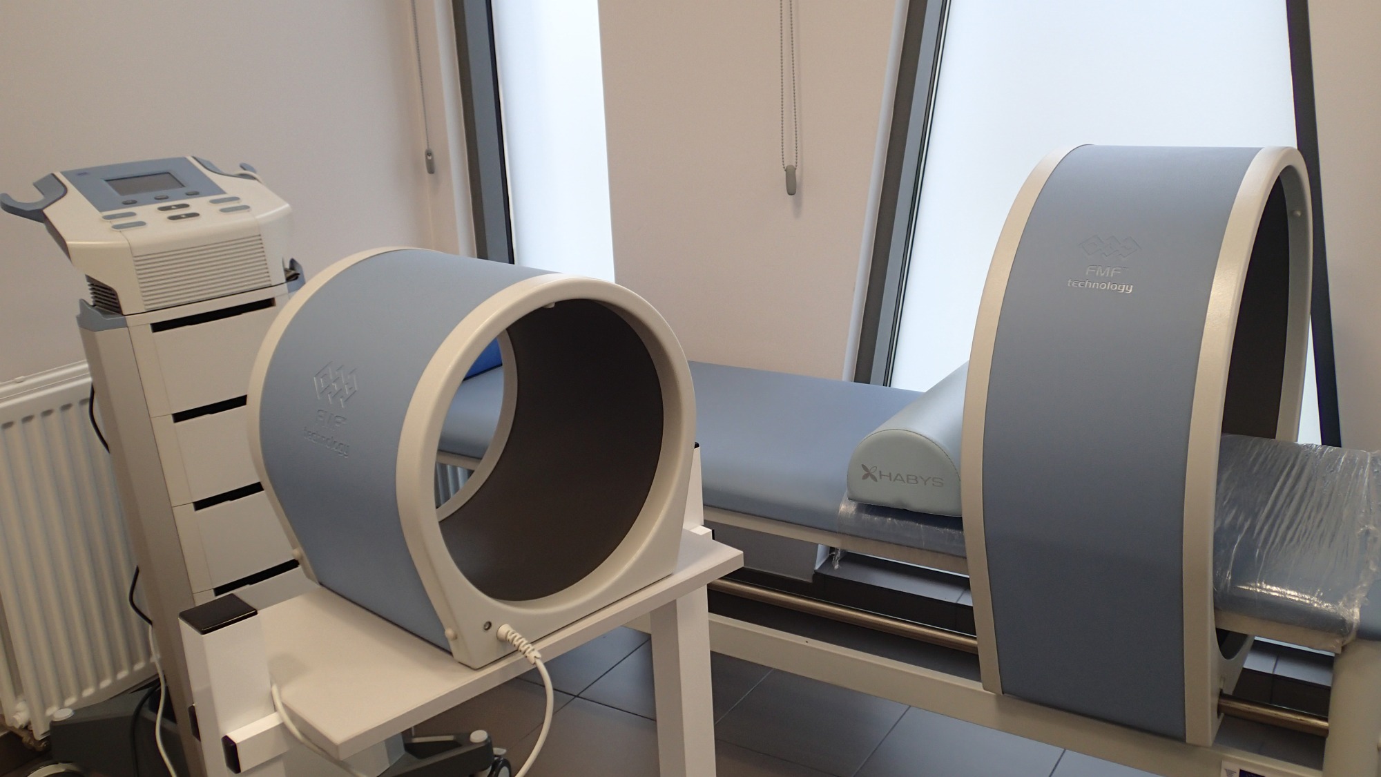 Aparat do magnetoterapii FMF Technology na tle jasnego wnętrza gabinetu w Dziwnowie, z widocznym łóżkiem rehabilitacyjnym i urządzeniem sterującym.