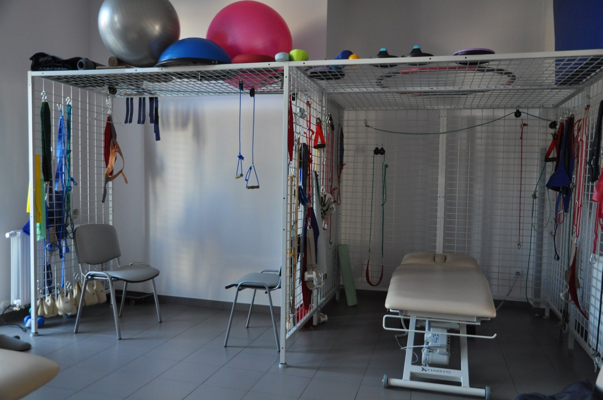 Sala rehabilitacyjna z drabinkami, stołem do masażu i piłkami do ćwiczeń, widoczne różnorodne akcesoria do rehabilitacji zawieszone na drabinkach.
