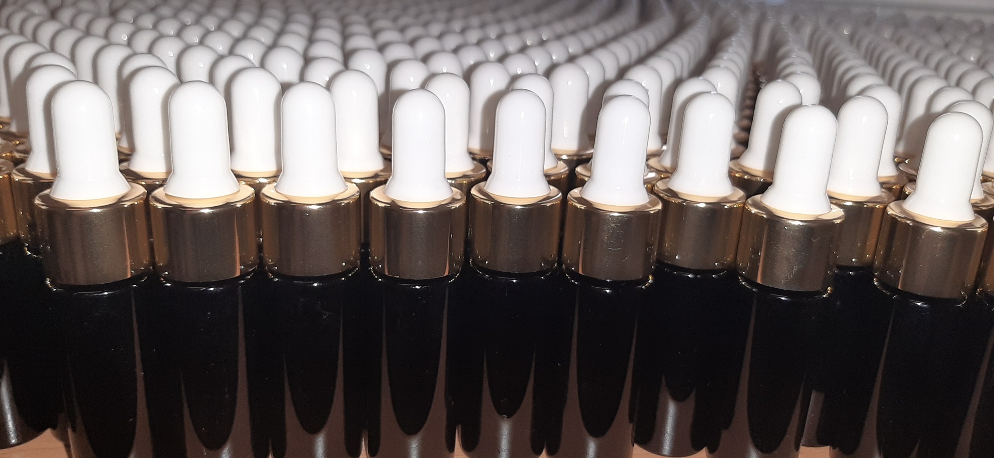 Rząd czarnych buteleczek z białymi pipetami i złotymi nakrętkami, ustawionych blisko siebie, prawdopodobnie serum lub olejki do pielęgnacji paznokci lub skórek.