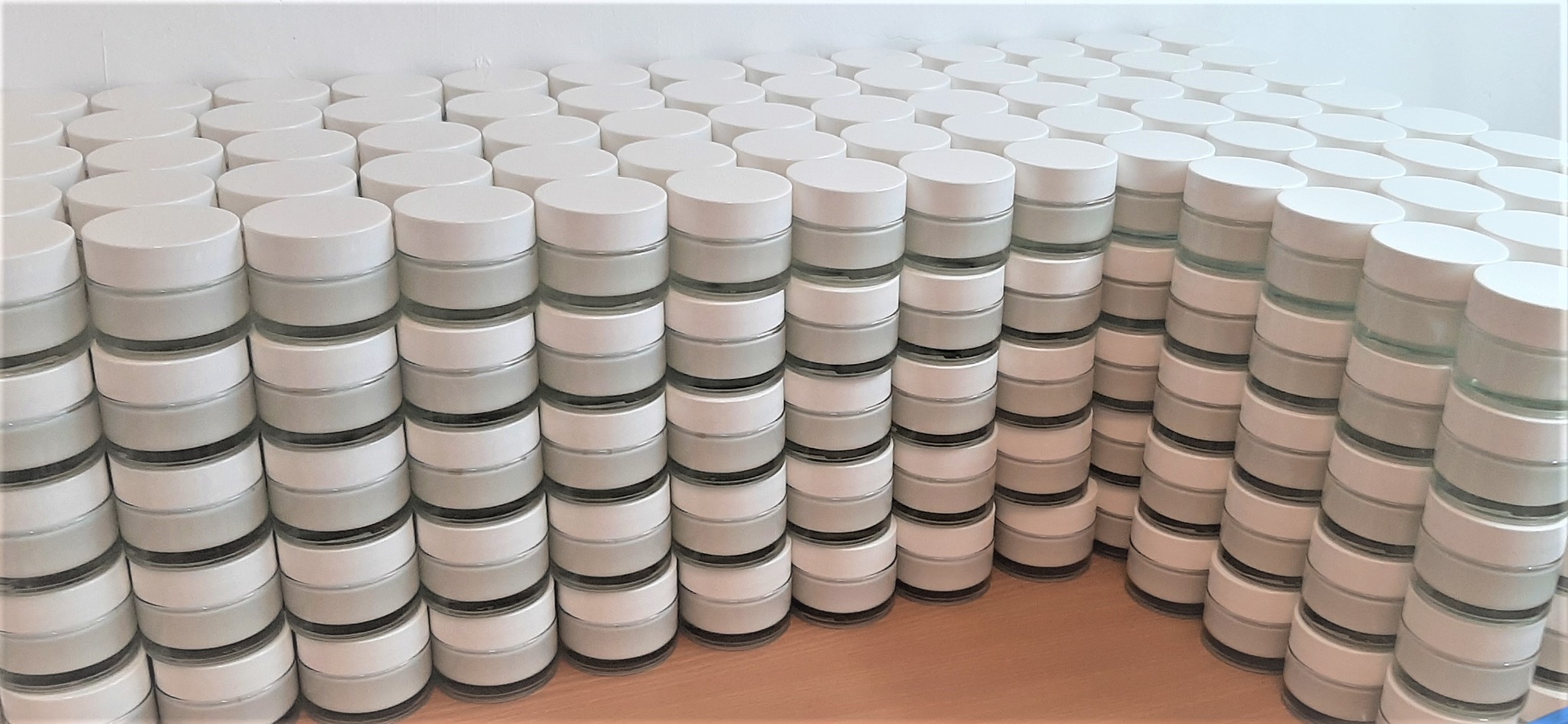 Rzędy identycznych, przezroczystych słoiczków z białymi wieczkami, wypełnionych kremem o neutralnym odcieniu, ustawione na drewnianej powierzchni.