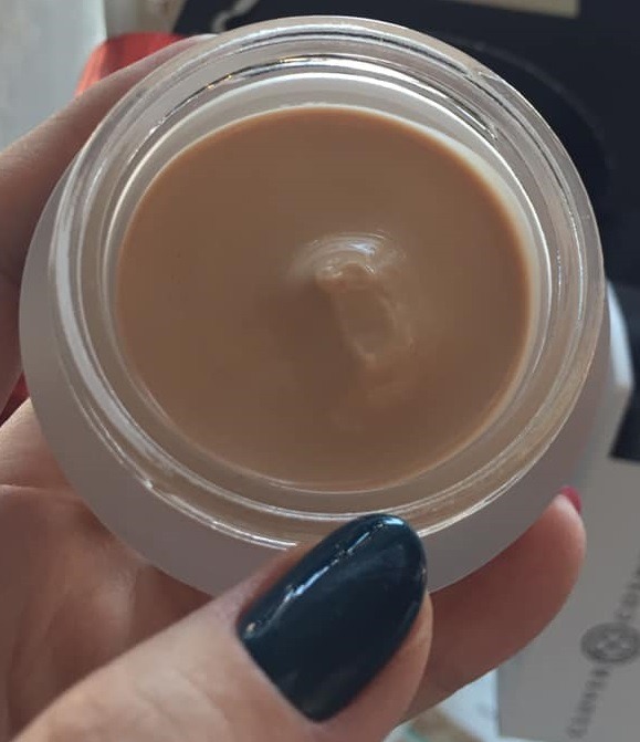 Otwarty słoiczek beżowego kremu do rąk trzymany w dłoni z pomalowanym na ciemnozielono paznokciem, widoczne logo producenta na opakowaniu w tle.