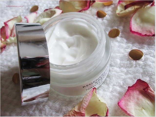 Otwarty słoiczek z białym kremem na ręczniku, otoczony płatkami róż i złotymi cekinami, widoczne odbicie światła na srebrnej pokrywce.