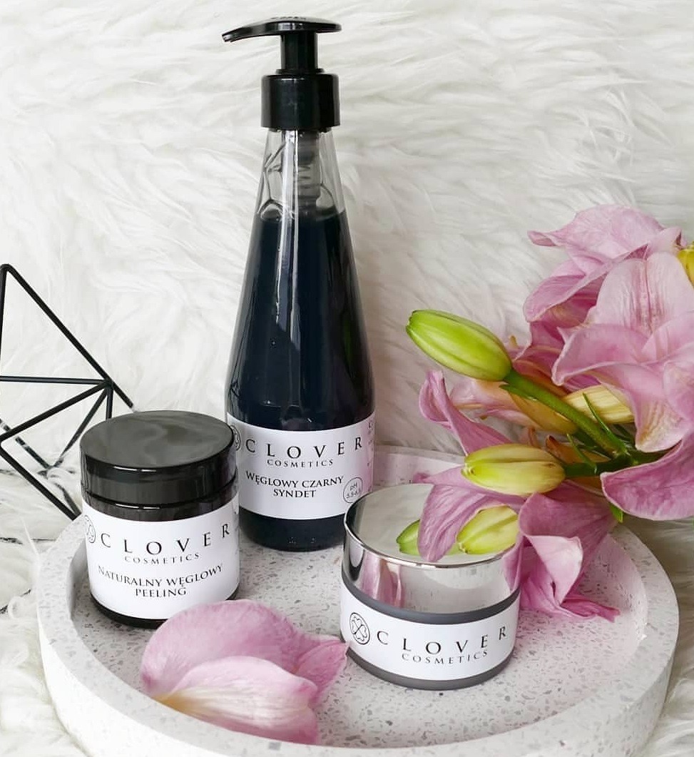 Zestaw kosmetyków Clover Cosmetics na betonowej tacy z płatkiem kwiatu i różowymi liliami, w skład którego wchodzą: naturalny węglowy peeling, węglowy czarny syndet oraz krem do pielęgnacji.