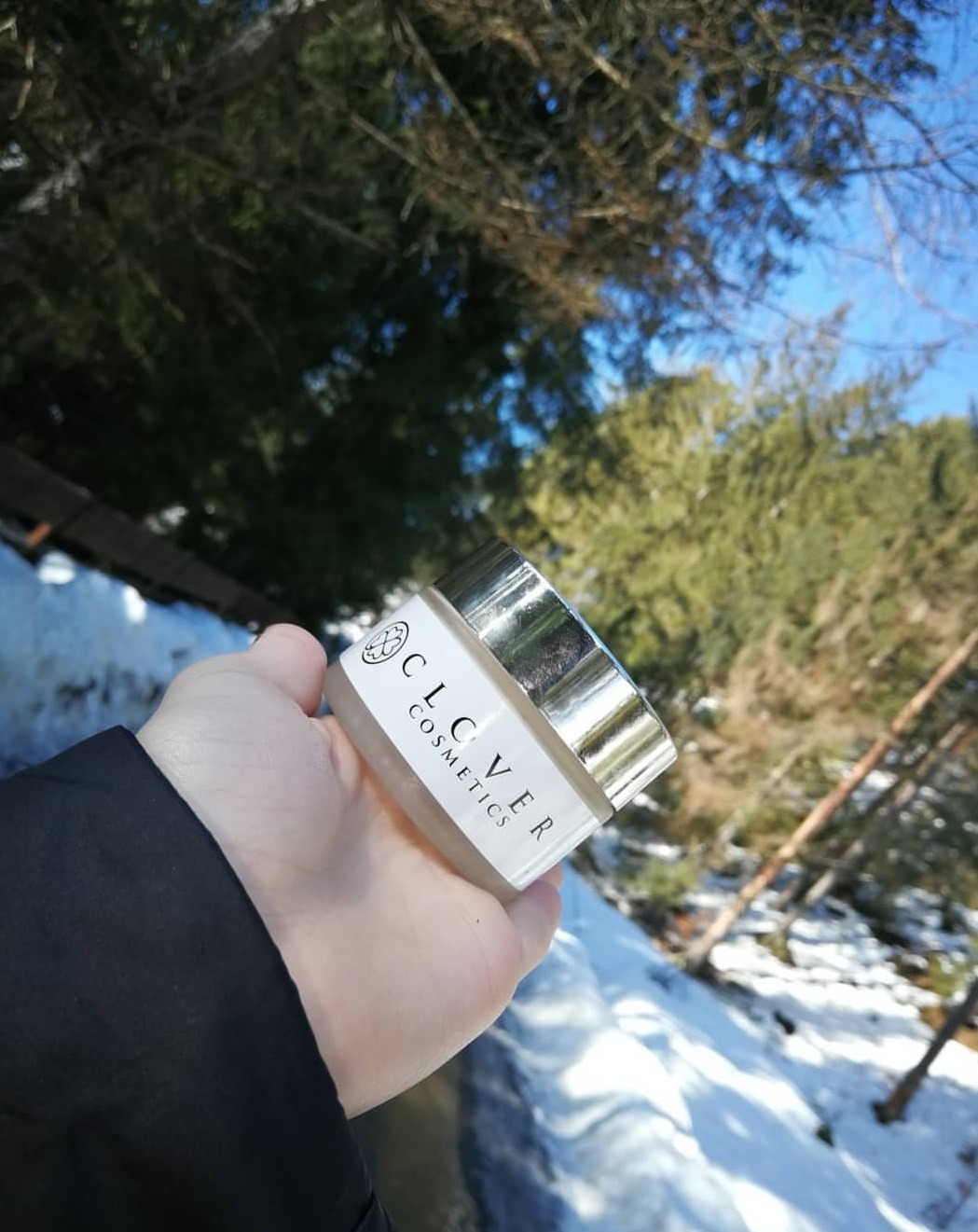 Osoba trzyma słoiczek kremu CLOVER Cosmetics na tle zimowego krajobrazu z drzewami i śniegiem.