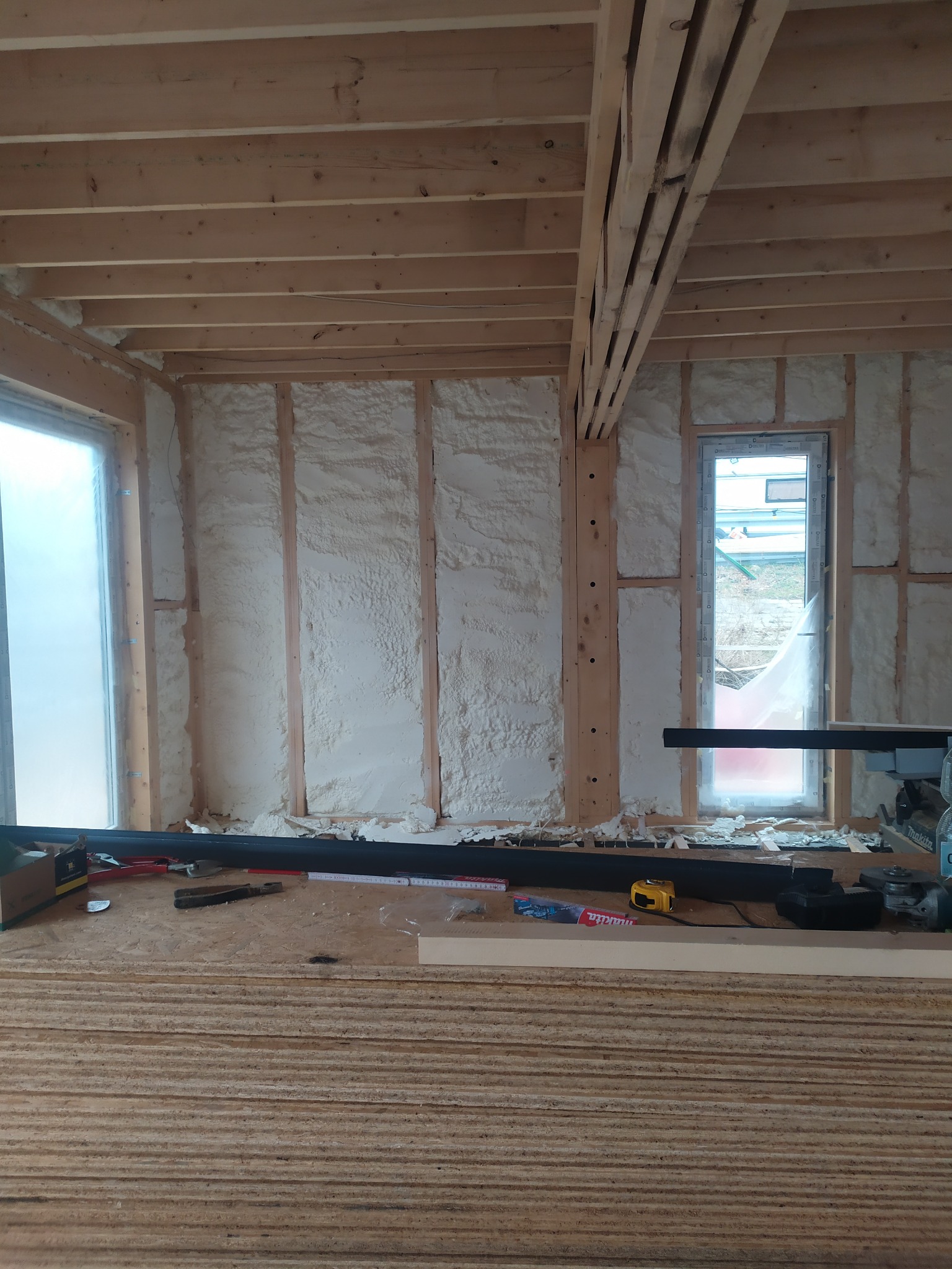 Wnętrze budynku w trakcie ocieplania pianką PUR, widoczne drewniane belki konstrukcyjne i narzędzia na podłodze, okna zabezpieczone folią.