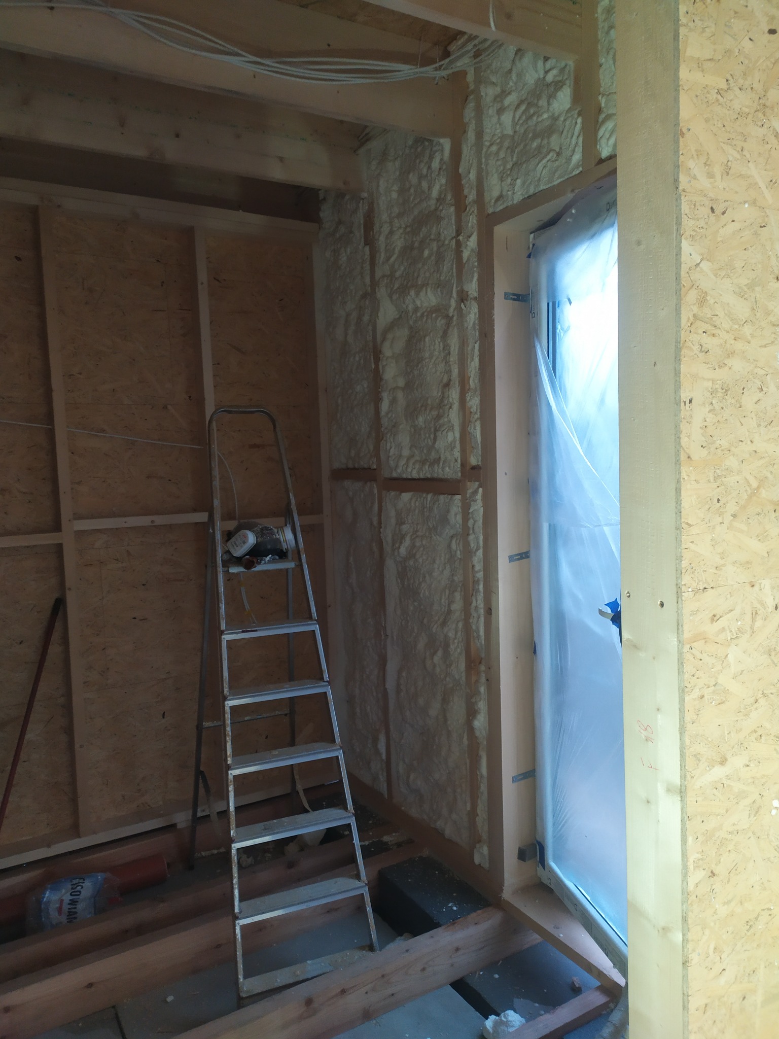 Poddasze w trakcie ocieplania pianką: widoczna struktura pianki PUR na ścianie, zabezpieczone folią okno, drabina aluminiowa i elementy konstrukcji drewnianej.