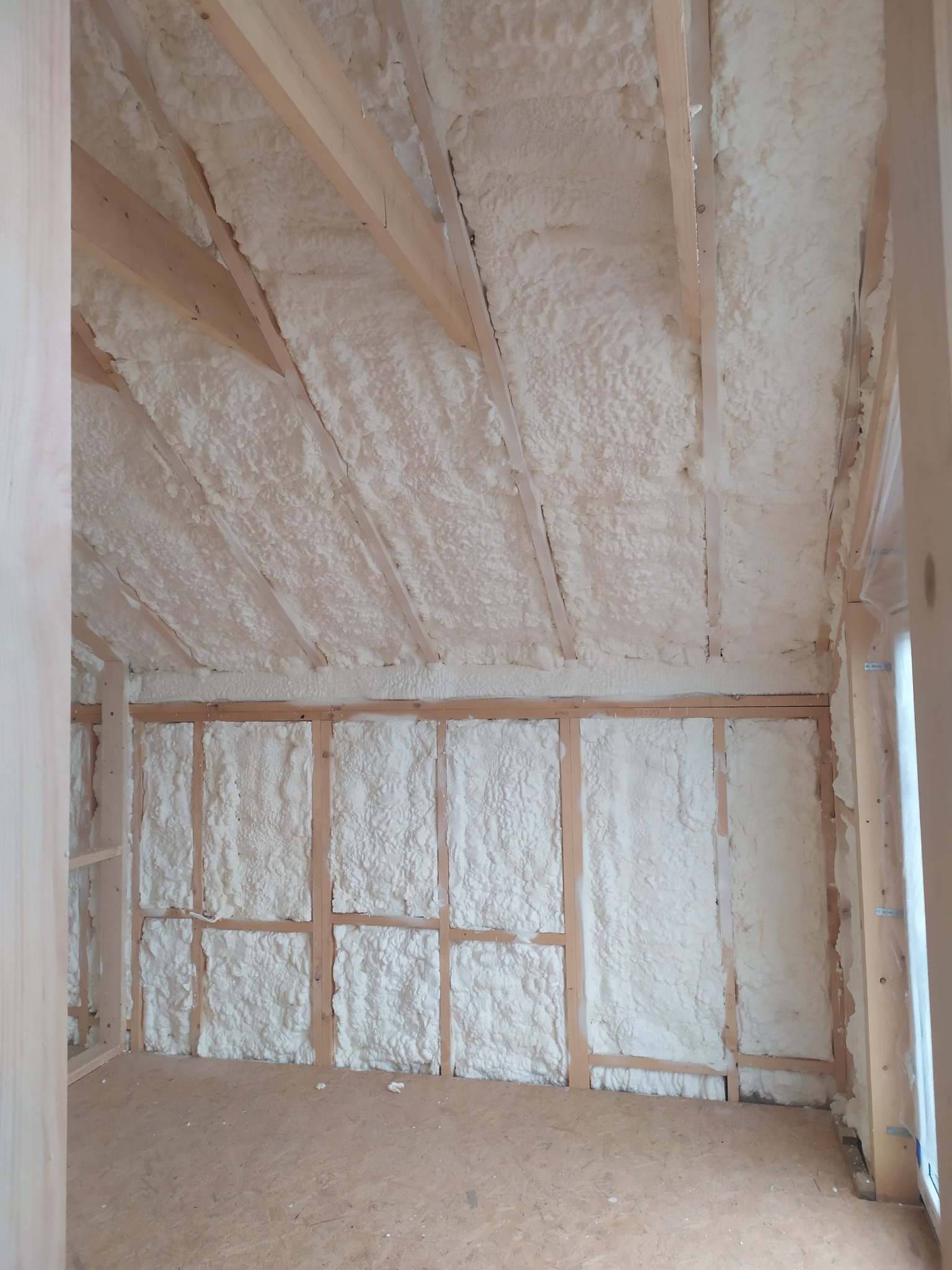 Poddasze po ociepleniu pianką PUR, widoczne drewniane krokwie i konstrukcja ścian wypełniona białą pianą, surowa podłoga z płyt OSB.